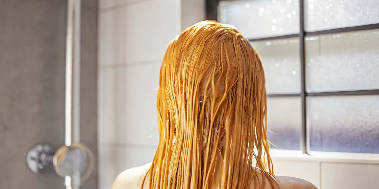 Woman gently rinsing conditioner through glossy color-treated brown hair in a bright minimalist bathroom